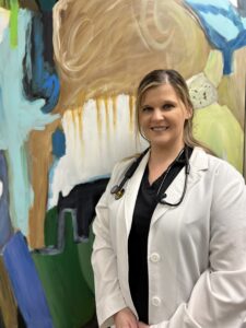 A woman in white lab coat standing next to a painting.