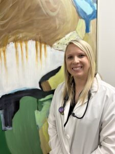 A woman in white lab coat standing next to a painting.